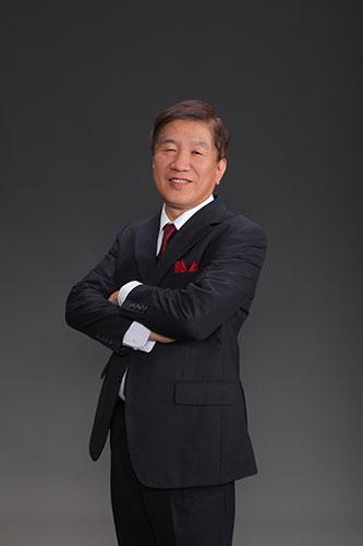 Smiling Asian man in a dark suit with a red pocket square stands with arms crossed against a gray studio backdrop.