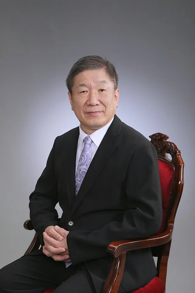 Portrait of a middle‑aged Asian man in a black suit and lavender tie, seated in a carved wooden chair against a gray studio backdrop.