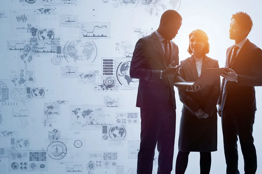 Three professionals in business attire discuss a tablet, with a blue backdrop of digital charts and world maps.